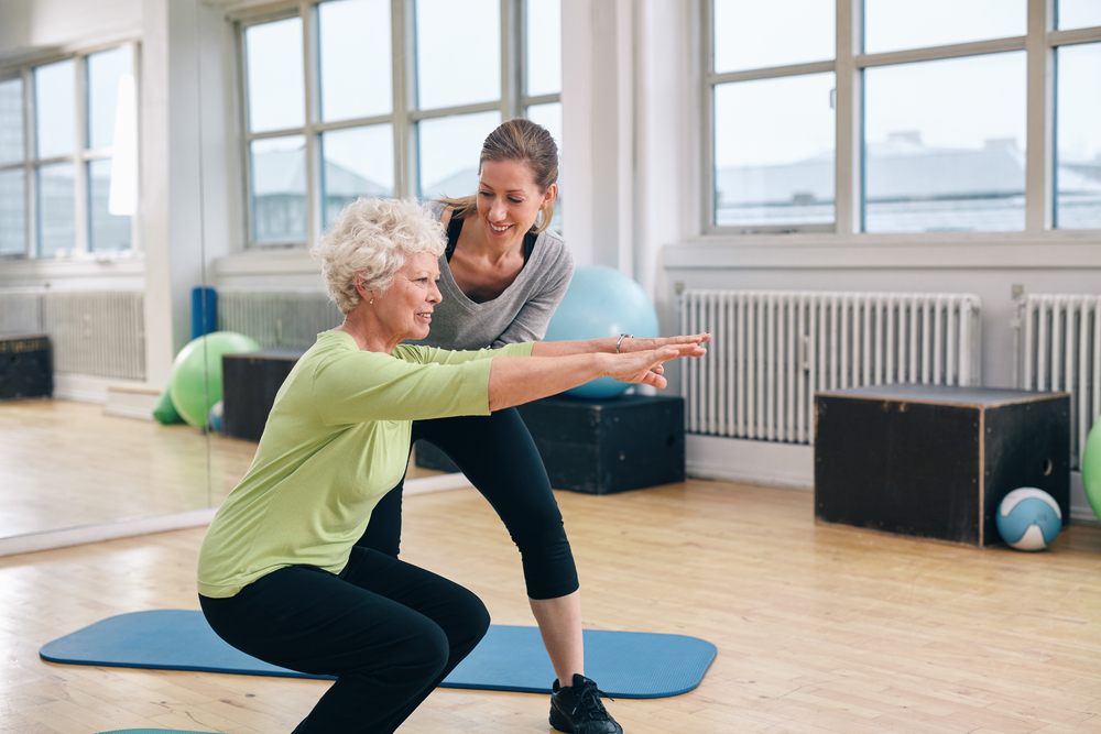 an older woman in a green shirt receives assistance from her physical therapist doing squats for pelvic floor therapy - highlighting the different types of physical therapy. 