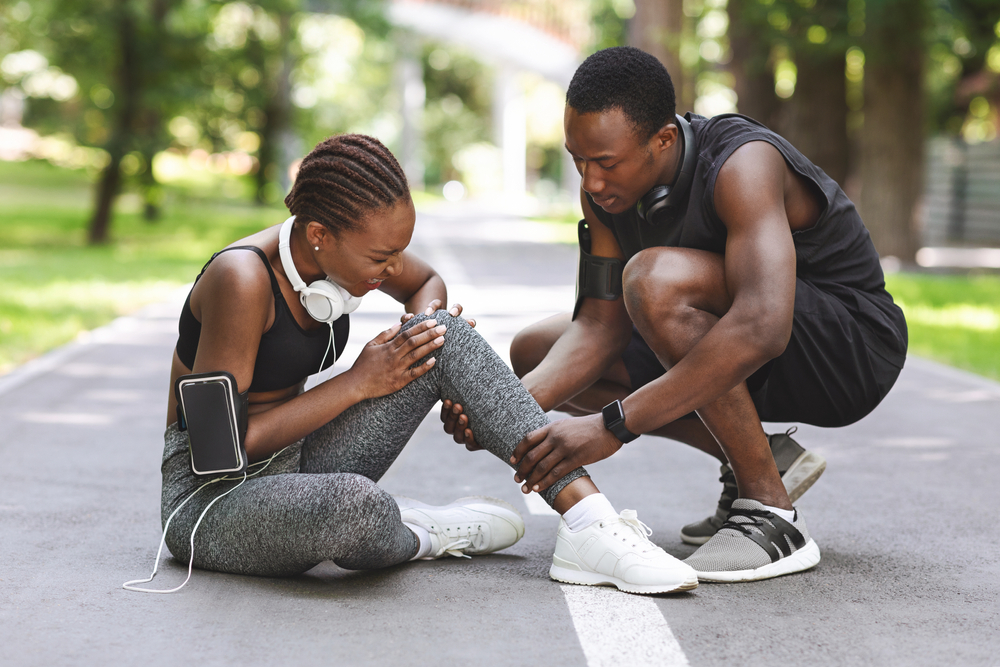 Woman and her running partner stop in the middle of the path, the woman holds her knee in pain while her running partner looks concerned