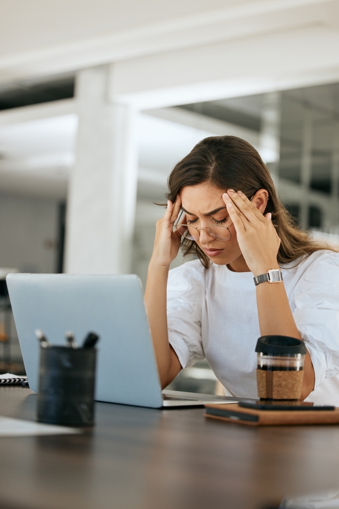 Woman at work, working off a laptop, rubbing her temples, she has a headache, her eyes are closed in pain